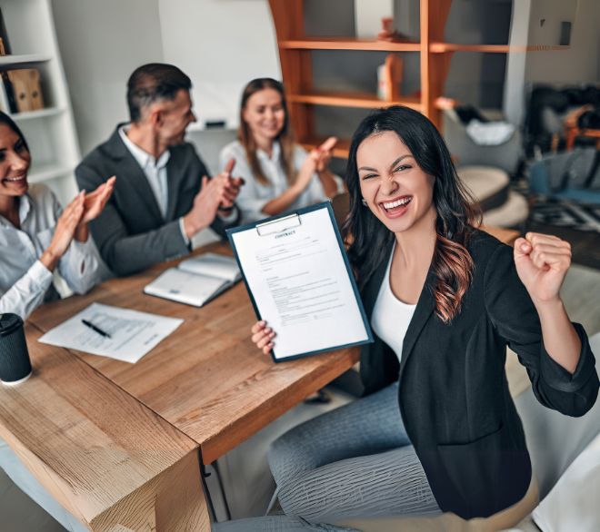 A woman celebrates receiving a job offer, holding a contract with excitement as a team applauds her success