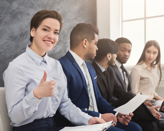 A confident candidate gives a thumbs-up while waiting for a job interview, sitting among other professionally dressed applicants